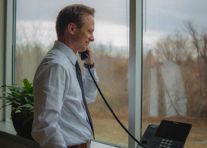 a man in a white button shirt talking on a phone with a window of his office behind him